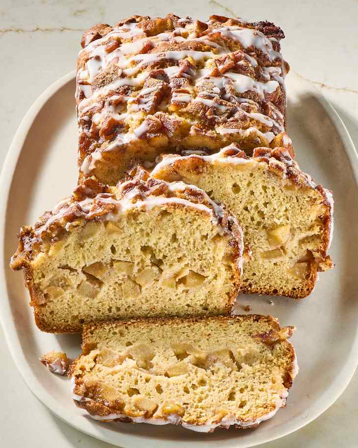 Sliced apple bread loaf drizzled with icing, showcasing apple chunks inside, on a beige plate.